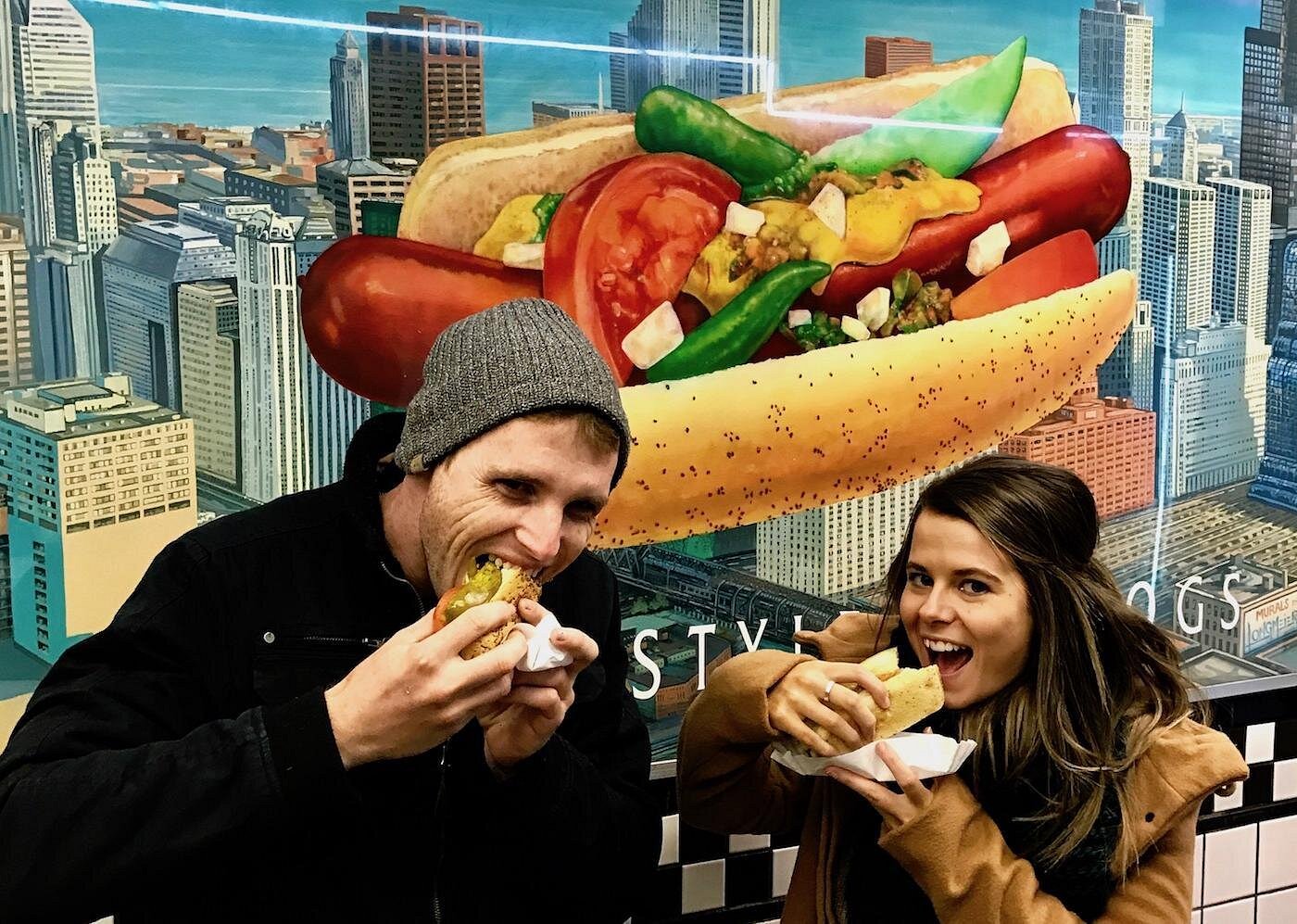 Two people eating hot dogs in front of a large hot dog mural in a city setting.